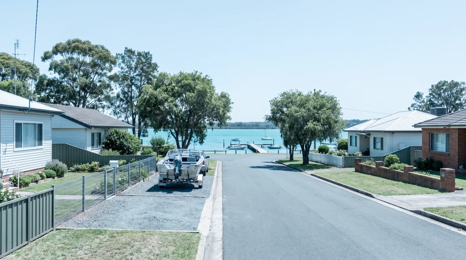 An established Belmont street on the eastern shore of Lake Macquarie at midday: single-storey weatherboard and brick homes on timber floors, a small aluminium boat on a trailer in a driveway, mature shade trees, and a glimpse of the blue-green lake and a jetty at the end of the street