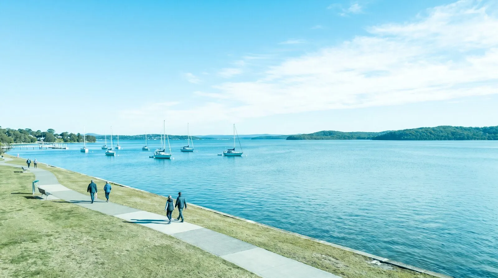 A wide midday view across Lake Macquarie from a grassy lakeside esplanade, flat blue-green water, moored yachts and low green headlands beyond