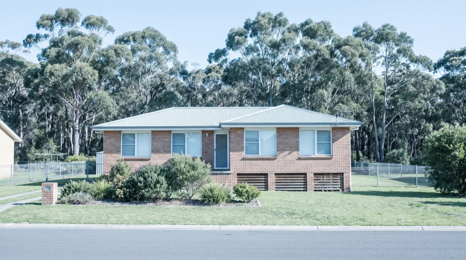 A brick Morisset home on low piers with a timber subfloor vent, tall grey-green eucalyptus bushland rising directly behind the back boundary at midday