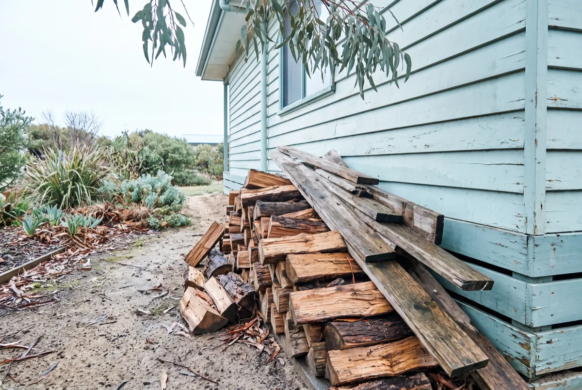 A stack of split firewood and stored timber piled directly against the weatherboard wall of a Morisset home near the ground, eucalyptus branches overhead