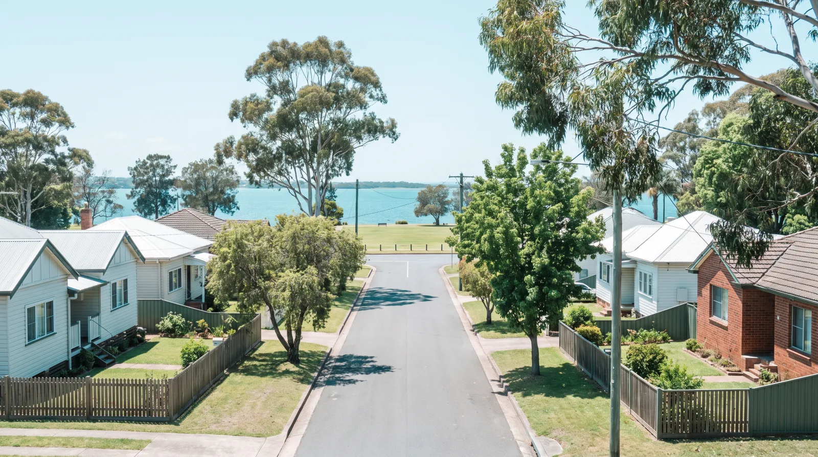 An established Speers Point street at midday running down to the lake: single-storey weatherboard and brick homes on timber floors, mature gum and shade trees, low front fences, a grassy foreshore reserve and blue-green lake at the end of the road